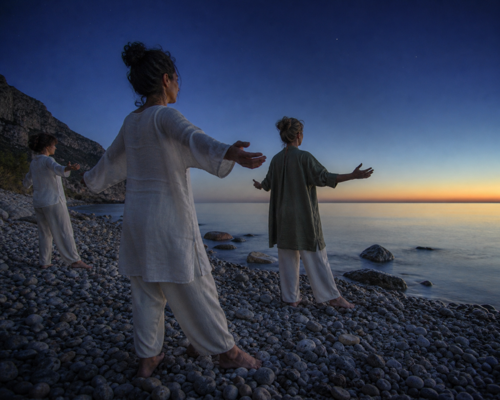 Three women practice sunrise yoga on a pebble beach along the Ionian coast of Calabria, Italy, as the sky shifts from navy to amber at the water's edge.